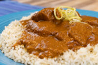 Butter Chicken pieces in thick brown sauce over rice on a blue plate, topped with yellow shredded garnish.