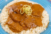 Butter Chicken pieces in thick orange sauce over rice, topped with shredded garnish, on a blue plate.