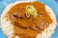 Butter Chicken pieces in reddish-brown sauce over white rice, topped with thin julienned garnish, on a blue plate