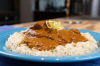 Butter Chicken in a brown tomato sauce over white rice on a blue plate, garnished with citrus zest.