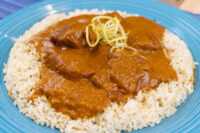 Butter Chicken pieces in thick tomato sauce over herbed rice, garnished with citrus zest, on a blue plate.