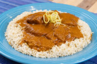 Butter Chicken pieces in tomato sauce over herbed rice on a blue plate, garnished with julienned zest.