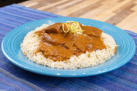 Blue plate of Butter Chicken over rice, garnished with citrus zest curls, on a striped blue cloth