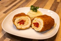 Sliced breaded Chicken Cordon Bleu roll with pimento pepper filling, plated with piped mashed potato and parsley.