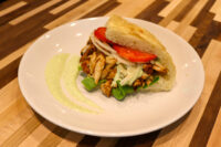 Chicken Gyros sandwich with grilled chicken strips, lettuce, onion, and tomato in flatbread, plated with green sauce.