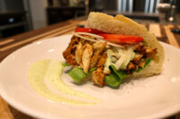 Chicken Gyros sandwich with sliced chicken, tomato, onion, spinach and green sauce in a roll on a white plate.