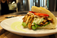 Chicken Gyros sandwich with grilled chicken strips, lettuce, tomato and onion in a bread roll on a white plate.