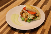 Chicken Gyros sandwich with grilled sliced chicken, lettuce, tomato and onion in a roll, green sauce swirl on plate.