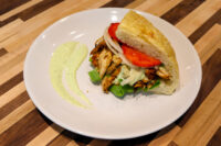 Chicken Gyros in flatbread with lettuce, onion, and tomato, served with a green herb sauce on a white plate.