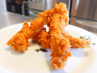 Four crispy Chicken Tenders stacked on a white plate with a small herb sprig garnish.