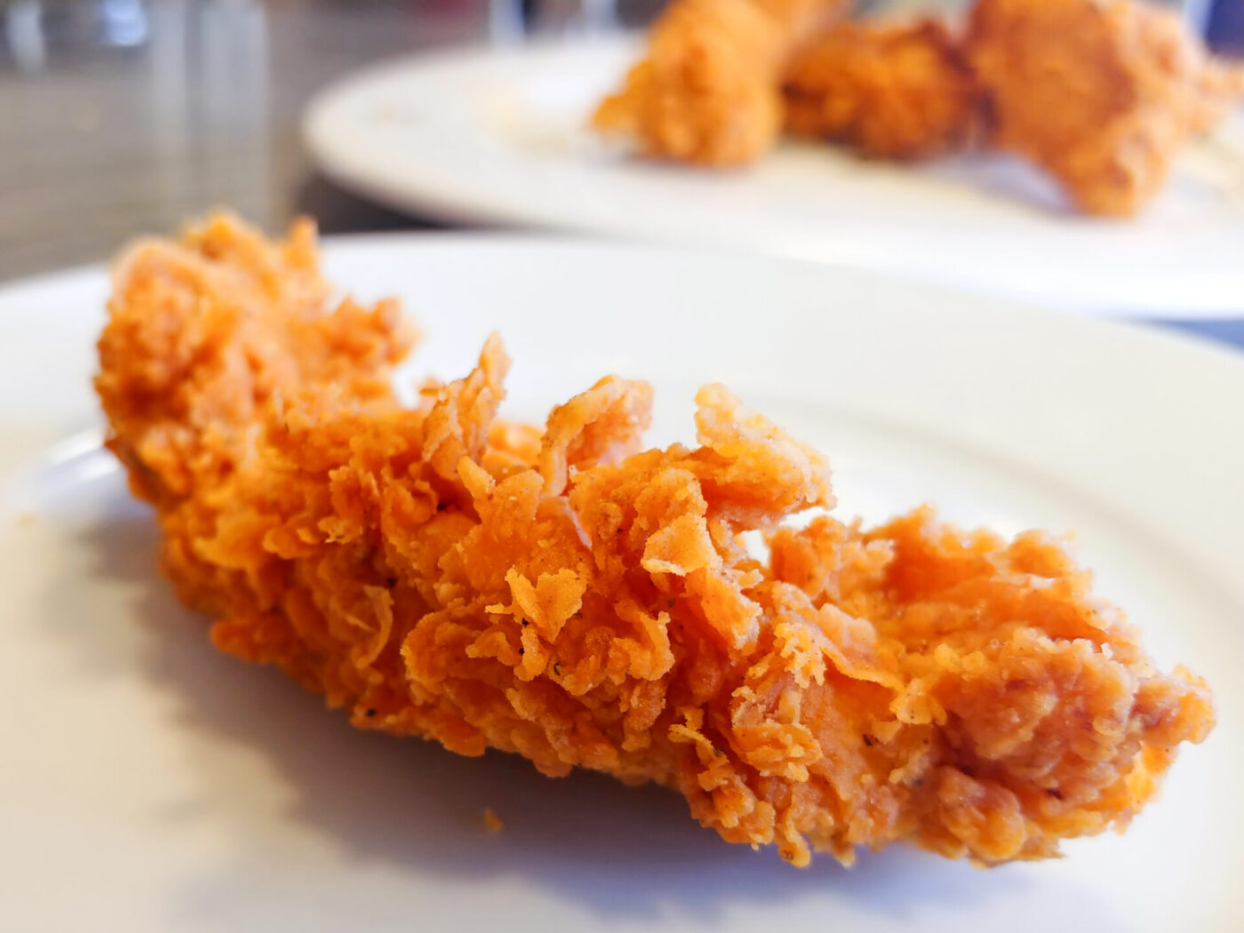Close-up of a crispy, breaded Chicken Tender on a white plate, more tenders visible on a plate behind.
