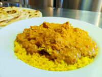Chicken Tikka Masala pieces in creamy orange sauce over yellow rice, naan bread visible behind the plate.