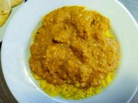 Chicken Tikka Masala in a white bowl, its creamy orange sauce covering turmeric-yellow rice, with a piece of flatbread beside it