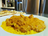 Chicken Tikka Masala in creamy orange sauce over yellow rice, with naan bread visible behind the plate.