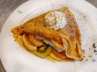Folded Crepes filled with banana, orange, kiwi and apple slices, topped with whipped cream and a dusting of spice on a white plate.
