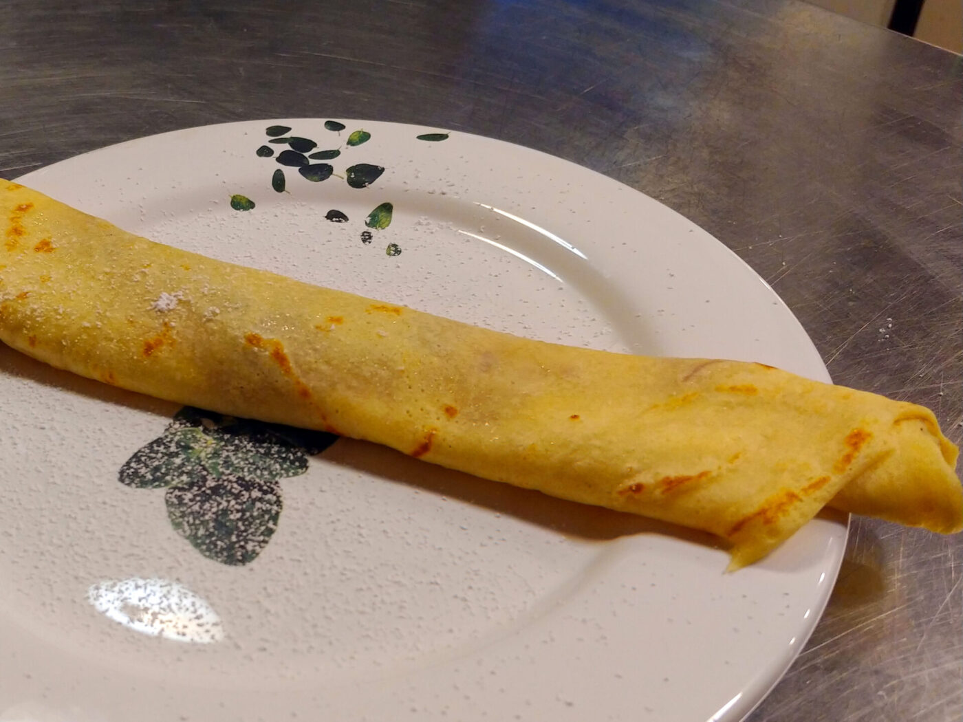 A rolled Crepes with a lightly speckled golden surface rests diagonally on a white patterned plate.