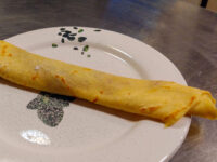 A rolled Crepes with a lightly speckled golden surface rests diagonally on a white patterned plate.