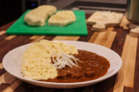 Plate of Czech Beef Goulash with sliced onions and a bread dumpling, more dumplings on a board behind