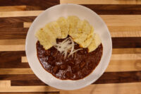 Bowl of Czech Beef Goulash with meat chunks, sliced onion, and grilled bread on a striped wood table.