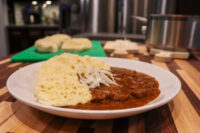 Plate of Czech Beef Goulash with paprika gravy, raw onion strips, and a scoop of pale dumpling.