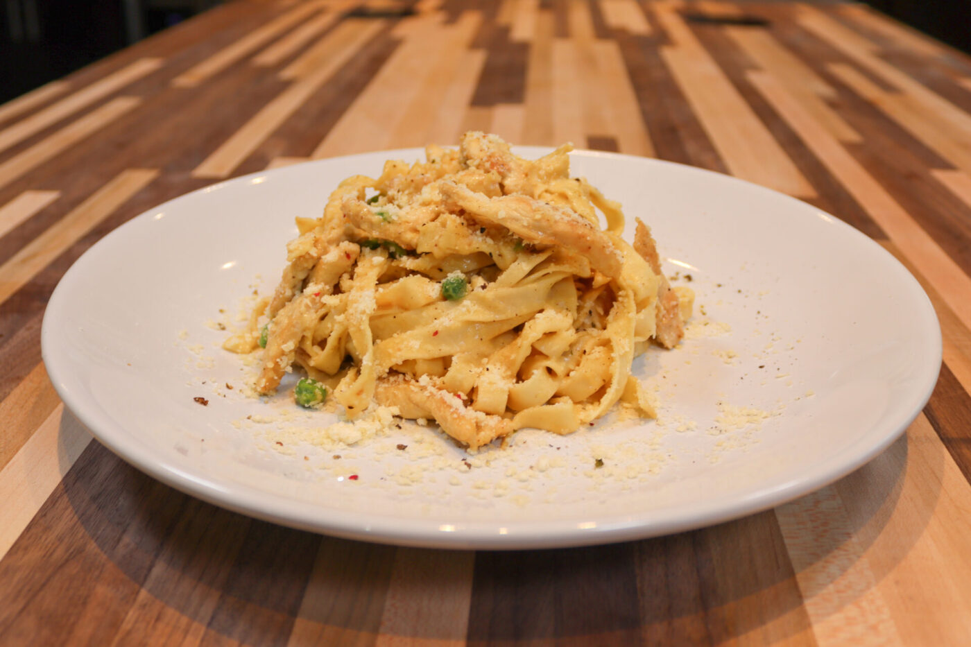 Fettuccine Alfredo with Chicken piled on a white plate, showing cream-coated ribbon pasta, chicken strips, peas, and grated cheese on a striped wooden table.