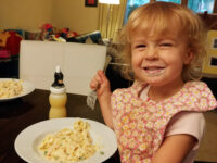 Toddler with sauce on her face holds a fork over a plate of Fettuccine Alfredo with peas in creamy sauce.