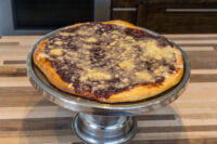 Whole Frgal Kolach with Plum Filling, dark filling and streusel visible, on a silver pedestal stand.
