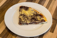 Triangular slice of Frgal Kolach with Plum Filling topped with streusel, on a white plate.