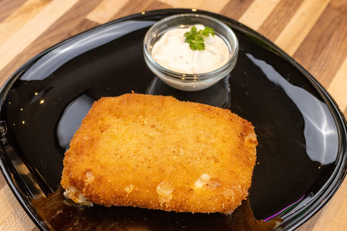 Golden breaded Fried Cheese cutlet on a black plate beside a small dish of herbed white dip.