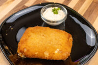 Golden breaded Fried Cheese cutlet on a black plate beside a small dish of herbed white dip.