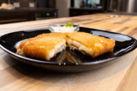 Fried Cheese with Ham cut in half showing melted cheese and ham, served with a small dip on a black plate.