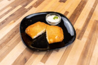 Two breaded, fried halves of Fried Cheese with Ham on a black plate, with a creamy dip and herb garnish.