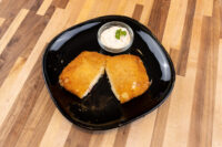 Two halves of Fried Cheese with Ham, breaded and golden, on a black plate with a creamy herb-topped dip.