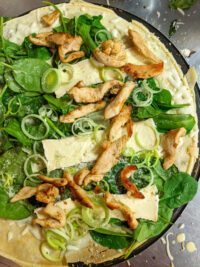 Overhead view of Galettes with Chicken, Spinach, and Brie Cheese, with curled leeks and grated cheese scattered over the top on a metal surface.
