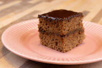 A square Gingerbread Bar with dark chocolate glaze, showing two spiced cake layers, on a pink plate.