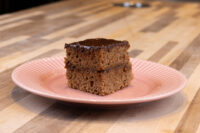 A square Gingerbread Bars slice with dark chocolate coating and a filling layer, on a pink ridged plate.