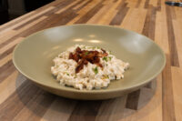 Bowl of Halušky with Bryndza Cheese Substitution topped with crispy bacon strips and herbs, on a wooden counter.