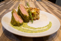 Sliced Herb-crusted Pork Tenderloin with pink center beside a potato bake and green herb sauce.