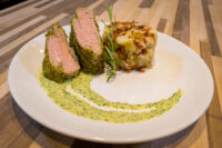 Sliced Herb-crusted Pork Tenderloin with pink centre beside a potato cake and green herb sauce.