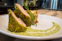 Sliced Herb-crusted Pork Tenderloin with pink center, green herb crust, rosemary garnish, and herb sauce swirl.