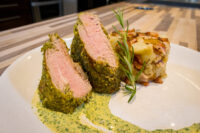 Sliced Herb-crusted Pork Tenderloin with green herb sauce beside a potato bacon bake and rosemary sprig.