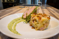 Sliced Herb-crusted Pork Tenderloin with rosemary, potato-bacon cake, and green herb sauce on a white plate.
