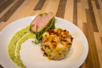 Sliced Herb-crusted Pork Tenderloin with rosemary, potato hash, and a green herb sauce swirl on a white plate.