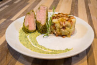Sliced Herb-crusted Pork Tenderloin with pink center, rosemary sprig, herb sauce, and potatoes on a white plate