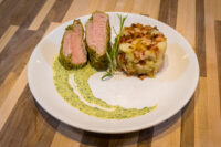 Sliced Herb-crusted Pork Tenderloin with pink centre, rosemary sprig, potato-bacon hash, and green herb sauce.