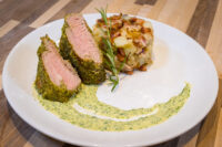 Sliced Herb-crusted Pork Tenderloin with pink centers beside roasted potatoes and herb sauce.
