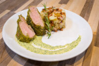 Sliced Herb-crusted Pork Tenderloin with pink center beside mashed potato and bacon on a swirl of green sauce