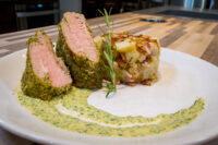 Sliced Herb-crusted Pork Tenderloin with pink centers beside a potato stack and green sauce on a white plate.