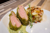 Sliced Herb-crusted Pork Tenderloin with pink center beside roasted potatoes and rosemary on a white plate.