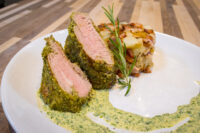 Sliced Herb-crusted Pork Tenderloin with pink center, roasted potatoes, rosemary, and green herb sauce on a white plate